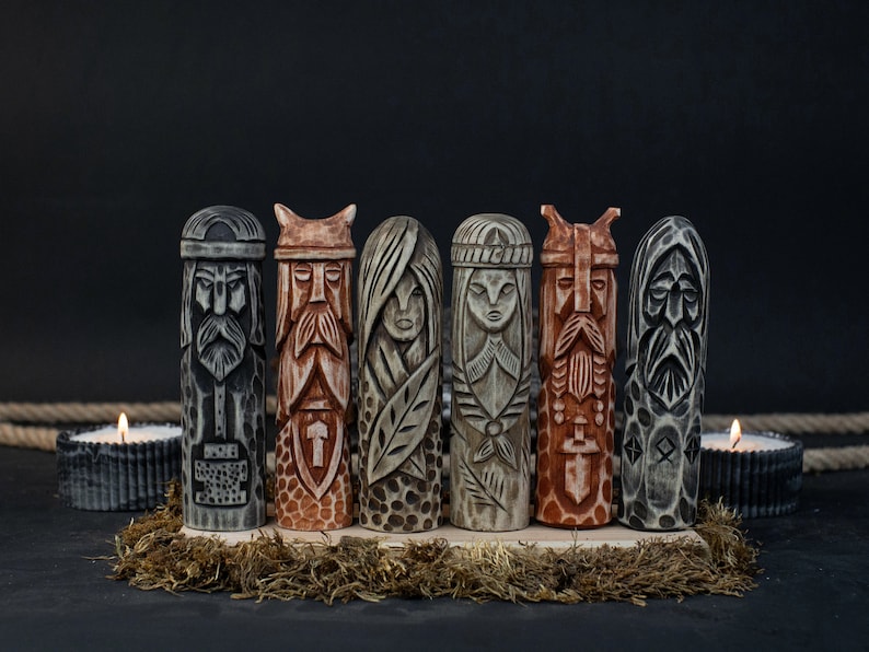 May include: Six carved wooden figures with detailed faces and intricate designs. The figures are in shades of gray, brown, and beige, and are arranged on a wooden base with moss. Two lit candles are in the background.