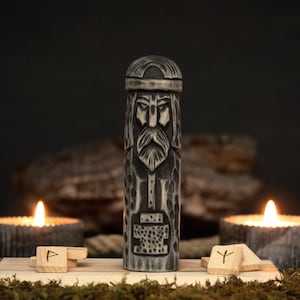 May include: A carved wooden statue of a bearded figure with a helmet, possibly a Viking god. The statue is gray and brown, with intricate details. Two lit candles and wooden runes are in the foreground, creating a mystical ambiance.