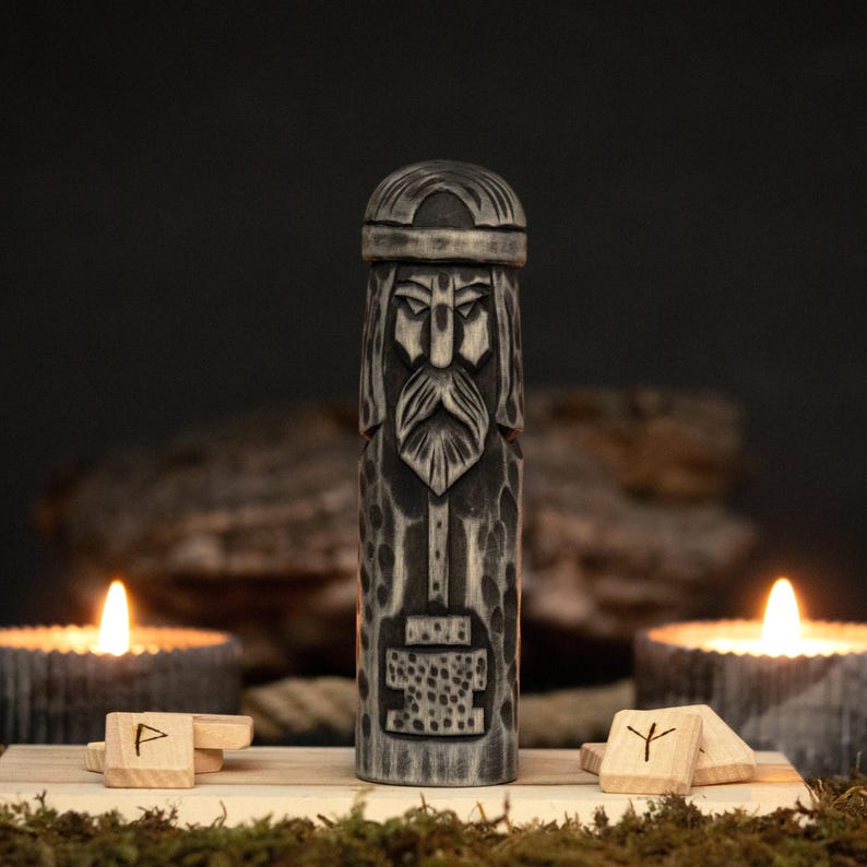 May include: A carved wooden statue of a bearded figure with a helmet, possibly a Viking god. The statue is gray and brown, with intricate details. Two lit candles and wooden runes are in the foreground, creating a mystical ambiance.