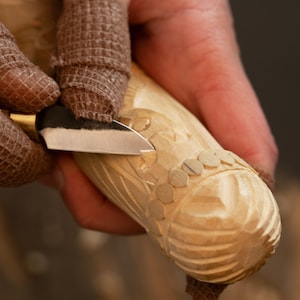 May include: Close-up of wood carving in progress. A hand, protected by a brown bandage, holds a carving knife, carefully shaping a light-colored wooden object. The object features carved circular and floral designs.