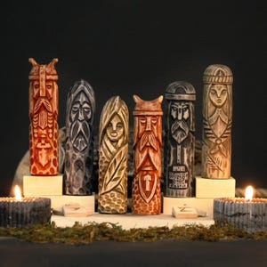 May include: Six carved wooden figures with detailed faces and intricate designs. The figures are in shades of brown, gray, and orange, each standing on a small wooden base. The background is dark, with candles and greenery adding to the ambiance.