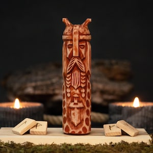 May include: A detailed wooden carving of a Viking figure with a helmet and long beard. The figure is standing on a wooden base with runes. Two lit candles and a blurred background complete the scene.