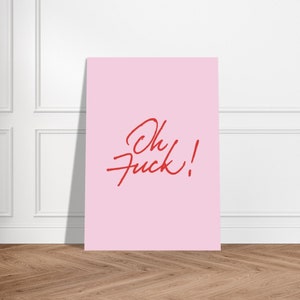 May include: A pink rectangular art print with the words "Oh Fuck!" in red cursive script. The print is leaning against a white wall and set on a wooden floor.