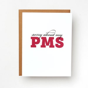 May include: A white card with a brown envelope. The card has black text that says "sorry about my PMS" in a cursive font. The word "PMS" is in large, bold, red letters.