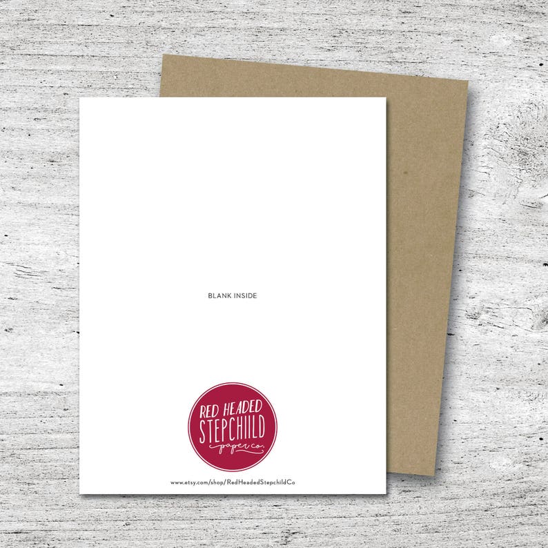 May include: A blank white greeting card with the text "BLANK INSIDE" and a red logo that says "RED HEADED STEPCHILD paperie". The card is paired with a brown envelope, set against a light wood grain background.