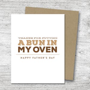 May include: A white Father's Day card with a brown envelope. The card reads "THANKS FOR PUTTING A BUN IN MY OVEN" in brown letters, with "HAPPY FATHER'S DAY" below. The card is on a light wood surface.