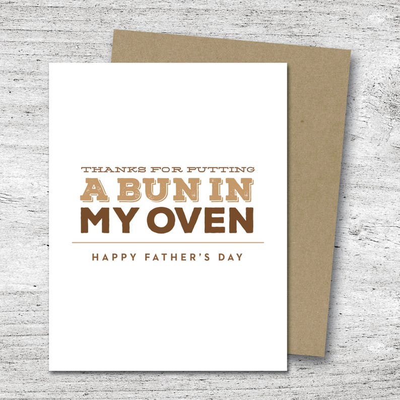 May include: A white Father's Day card with a brown envelope. The card reads "THANKS FOR PUTTING A BUN IN MY OVEN" in brown letters, with "HAPPY FATHER'S DAY" below. The card is on a light wood surface.