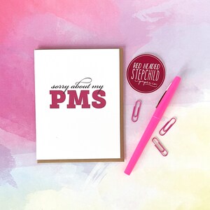 May include: A white card with the text "sorry about my PMS" in black and red lettering. The card is on a pink and yellow watercolor background with a pink pen and paper clips.