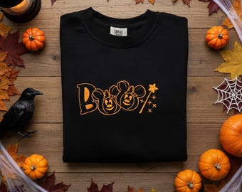 Peut inclure: Un sweat-shirt noir à col rond avec un motif brodé orange sur la poitrine. Le design affiche le mot Boo avec deux personnages en forme de citrouille rappelant des oreilles de souris et une petite baguette magique étoilée sur la droite.