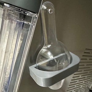 Op de afbeelding: Een transparante plastic ijsschep is bevestigd aan een grijze plastic houder die is gemonteerd op een apparaat van roestvrij staal. De schep is ontworpen om ijs te bevatten en is met een kleine schroef aan de houder bevestigd.