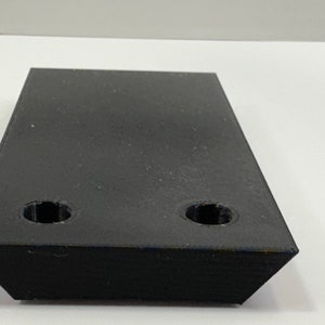 May include: A black rectangular block with two circular holes. The object has angled edges and appears to be made of a solid material. The item is set against a white background.
