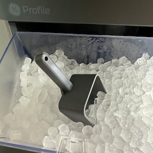 Op de afbeelding: Een doorzichtige plastic bak gevuld met ijsblokjes. Een grijze plastic ijsschep is gedeeltelijk ondergedompeld in het ijs.