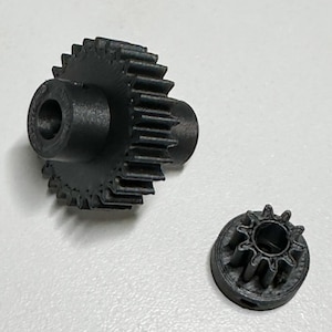 May include: Two black plastic gears, one larger with a central hole and a toothed outer edge, the other smaller and circular with teeth around the edge.