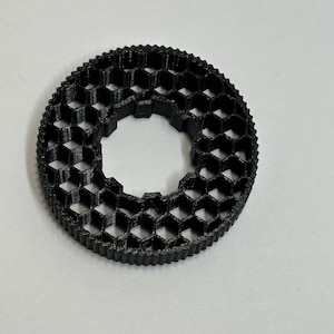 May include: A black, circular object with a honeycomb pattern. The object has a central hole and a ridged outer edge. The design is made of interconnected hexagonal shapes, creating a textured appearance. The object is likely 3D-printed.