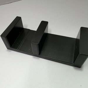 May include: A black, rectangular bookend with a minimalist design. The bookend features two upright supports connected by a flat base, creating a sturdy structure for holding books. The matte finish gives it a modern look.