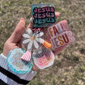 May include: A set of five resin keychains with glitter and colorful designs. The keychains feature the phrases "Jesus", "Fall in Love with Jesus", "God is Good", and "Jesus Loves You".