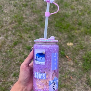 May include: Purple reusable tumbler with a straw and a bow on top. The tumbler has a glittery finish and features various stickers, including one that says "GOD'S" and another that says "LOVE".