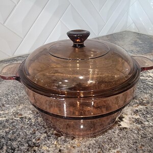 May include: A vintage brown glass casserole dish with a matching lid. The dish has two handles and a knob on the lid. The glass has a slight amber tint. The casserole dish is designed for cooking and serving.