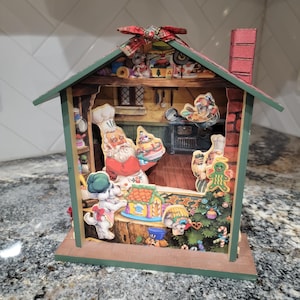 May include: A Christmas-themed diorama featuring Santa Claus and other characters in a kitchen setting. The house-shaped decoration has a green roof, red chimney, and a bow on top. Inside, Santa holds a tray of cookies.