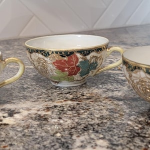 May include: Three white teacups with gold trim and floral designs. The cups have a black rim and feature red, green, and gold floral patterns.