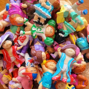 May include: A collection of small, colorful Cabbage Patch Kids figurines. The figurines are dressed in various outfits, including dresses, hats, and coats. Some hold accessories like hearts and purses. The colors include blue, pink, red, and yellow.