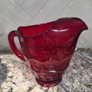 May include: A ruby red glass creamer with a handle and a spout. The creamer has a faceted design on the lower portion of the body. The handle is curved, and the spout is gently curved. The creamer is made of a transparent glass.