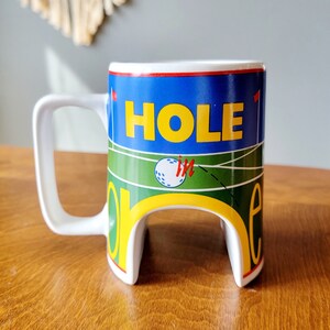 May include: White ceramic golf mug with a blue, green, yellow, and red design. The mug has the words "HOLE IN" printed on it.
