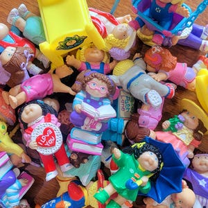 May include: A collection of Cabbage Patch Kids dolls in various poses and outfits. The dolls are made of plastic and have painted faces. Some of the dolls are holding accessories, such as books, umbrellas, and a heart-shaped sign that says "I love you".