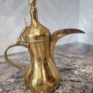 May include: A gold-colored, ornate coffee pot with a long, curved spout and a handle. The pot has a tall, slender neck and a decorative finial on the lid. The pot is made of metal and has a polished finish. The coffee pot is likely used for serving coffee.