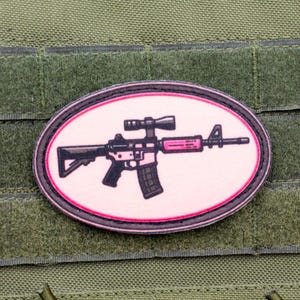 May include: A pink oval-shaped patch with a black and white illustration of a rifle with a scope. The patch has a black border.