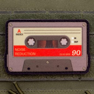 Pode incluir: Um patch retangular com um design de fita cassete retrô. A fita tem um rótulo vermelho com "NOISE REDUCTION" e "90" impressos. O patch é cinza, com detalhes em preto e está sobre um fundo verde.