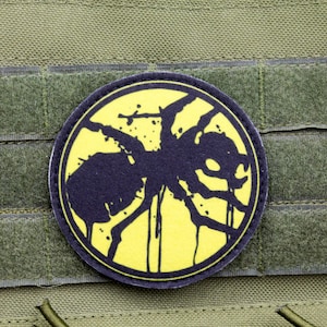 May include: A black and yellow patch with a stylized ant design. The patch is circular and has a black border. The ant is in the center of the patch and is facing to the right. The ant is outlined in black and has a yellow background.