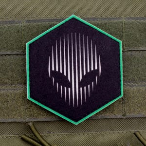 Pode incluir: Um patch hexagonal preto e verde com um design de rosto alienígena branco. O patch tem uma borda verde e é preso a um fundo de tecido verde com tiras de velcro.