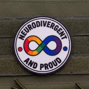 Peut inclure: Un écusson circulaire avec un fond blanc et une bordure noire. Il présente un symbole de l'infini aux couleurs de l'arc-en-ciel au centre. Le texte "NEURODIVERGENT" est arqué au-dessus et "AND PROUD" en dessous. Un point rouge et un bleu sont sur les côtés.