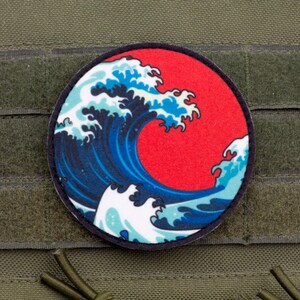 May include: A round patch featuring a stylized depiction of a large ocean wave in shades of blue and white against a red background. The patch is set against a green fabric background with hook and loop fasteners.