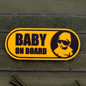Peut inclure: Un écusson jaune et noir avec les mots "BABY ON BOARD" et un bébé de dessin animé portant des lunettes de soleil. L'écusson est rectangulaire avec des coins arrondis et est posé sur un fond vert.