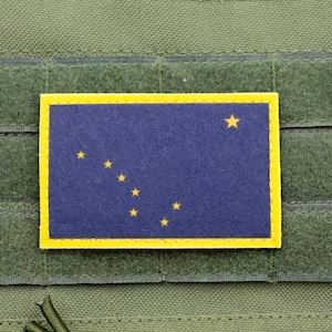 以下が含まれることがあります： アラスカ州旗をあしらった長方形のパッチ。濃い青色の背景に金色の星と黄色の縁取りが施されています。緑色の生地、おそらくタクティカルベストやバッグに取り付けられています。
