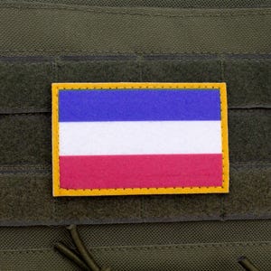 May include: A rectangular patch displaying the flag of Yugoslavia, with blue, white, and red horizontal stripes, edged in yellow. The patch is attached to a green fabric, possibly part of a tactical vest or similar equipment.