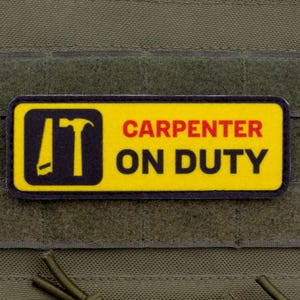 May include: A rectangular patch with a yellow background and black border. The patch features a hammer icon and the words "CARPENTER ON DUTY" in red and black. The patch is attached to a green fabric.