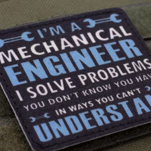 May include: A square patch with the text "I'M A MECHANICAL ENGINEER I SOLVE PROBLEMS YOU DON'T KNOW YOU HAVE IN WAYS YOU CAN'T UNDERSTAND" in white and blue on a black background. The patch is on a green fabric.