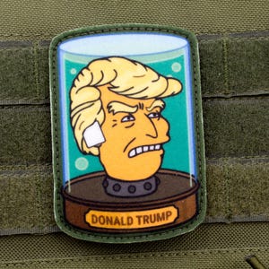 May include: A green and brown embroidered patch featuring a cartoon image of Donald Trump's head in a glass jar. The text "DONALD TRUMP" is printed at the bottom of the patch.