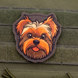 Puede incluir: Un parche colorido con la cara de un perro Yorkshire Terrier de dibujos animados con pelaje marrón, naranja y blanco, una nariz negra y una lengua rosa. El parche está adherido a un fondo de tela verde.