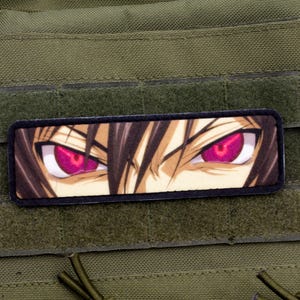 Op de afbeelding: Een rechthoekige patch met ogen in anime-stijl, magenta irissen en bruin haar. De patch is geplaatst tegen een groene achtergrond, waarschijnlijk een tactisch vest. De ogen hebben een vastberaden uitdrukking.