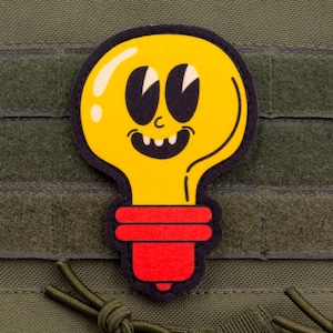 Op de afbeelding: Een gele lamp patch met een lachend gezicht, zwarte ogen en een rode basis. De patch zit op een groene stoffen achtergrond met klittenband en een trekkoord.