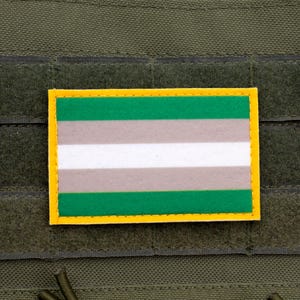 May include: A rectangular patch with a green, gray, and white striped design, outlined in yellow. The patch is affixed to a dark green fabric background, likely a tactical vest or similar gear.