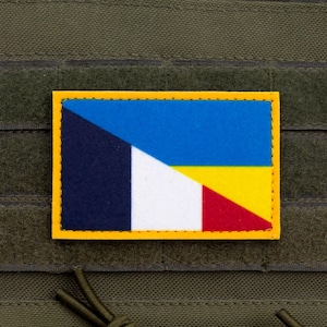 Peut inclure: Un écusson rectangulaire à bordure jaune, combinant les drapeaux de la France, de l'Ukraine et un élément bleu et blanc. L'écusson est fixé sur un fond en tissu vert.