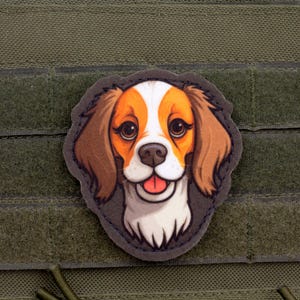Può includere: Toppa raffigurante un cane dei cartoni animati con pelo marrone e bianco, una lingua rosa e un'espressione felice. La toppa è su uno sfondo verde con una chiusura a strappo.