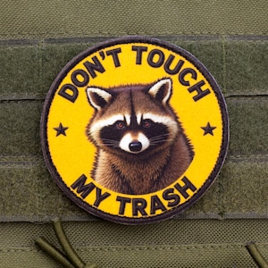 以下が含まれることがあります： 黄色い背景に「DON'T TOUCH MY TRASH」の文字とアライグマのイラストが描かれた丸いパッチ。アライグマの画像の両側に2つの黒い星があります。パッチは緑色の生地に取り付けられています。
