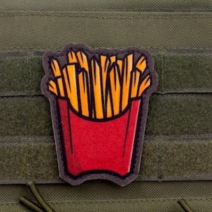 May include: A red patch featuring a cartoon depiction of a container of french fries. The fries are golden yellow and orange, and the container is red with a black outline. The patch is attached to a green fabric.