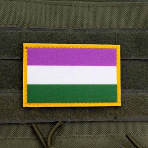 May include: A rectangular patch with a purple, white, and green striped design, framed by a yellow border. The patch is affixed to a dark green, textured fabric, possibly a tactical vest or similar item. The patch is a symbol of genderqueer pride.